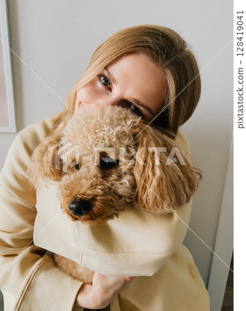 Woman with blond hair, dressed in casual clothes, lovingly embracing her small brown dog, radiating warmth and affection in cozy indoor setting, celebrating their bond. Blond woman hugging brown dog Woman with blond hair, dressed in casual clothes, lovingly embracing her small brown dog, radiating warmth and affection in cozy indoor setting, celebrating their bond. Blond woman hugging brown dog 128419041