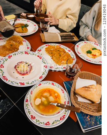 People are sharing a meal of potato pancakes, soup, salad, and bread, served on decorative plates and bowls in a cozy restaurant setting, highlighting the rich culinary traditions of Eastern Europe People are sharing a meal of potato pancakes, soup, salad, and bread, served on decorative plates and bowls in a cozy restaurant setting, highlighting the rich culinary traditions of Eastern Europe 128419048