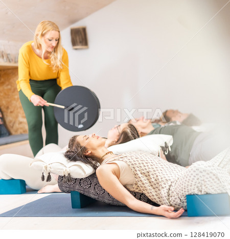 Group therapy session with Tibetan drum for group of women lying on wooden floor surrounded by copper bowls. Meditation and relaxation 128419070
