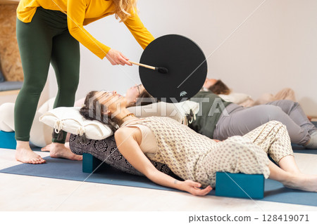 Group therapy session with Tibetan drum for group of women lying on wooden floor surrounded by copper bowls. Meditation and relaxation 128419071