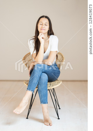 Portrait of confident beautiful woman with long brown hair, wearing casual clothes, sitting on chair in tight jeans and white t-shirt, studio background Portrait of confident beautiful woman with long brown hair, wearing casual clothes, sitting on chair in tight jeans and white t-shirt, studio background 128419073