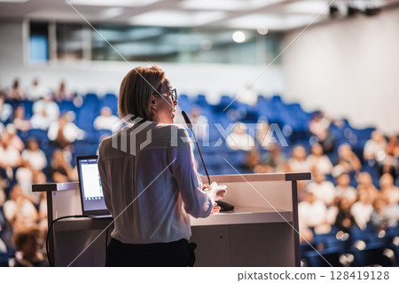 Female speaker giving a talk on corporate business conference. Unrecognizable people in audience at conference hall. Business and Entrepreneurship event 128419128