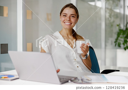 A happy and smiling professional sitting at their desk while enjoying coffee and using a laptop 128419236