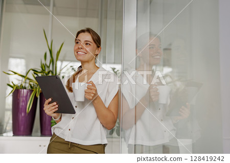 A young woman in a modern office holds a tablet and coffee, looking professional and thoughtful A young woman in a modern office holds a tablet and coffee, looking professional and thoughtful 128419242