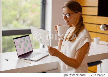 A woman is diligently working on her laptop surrounded by documents at her home office space 128419264