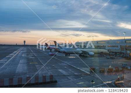 Airplanes from Etihad Airways and other airlines parked near a terminal with large windows, ground service vehicles, and a distant control tower at sunset. Airplanes from Etihad Airways and other airlines parked near a terminal with large windows, ground service vehicles, and a distant control tower at sunset. 128419420