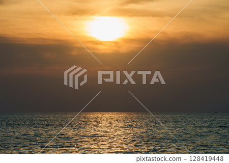 A tranquil sunset over the ocean, with the sun partially hidden by clouds. Warm golden light reflects on the calm sea, suggesting a tropical setting. A tranquil sunset over the ocean, with the sun partially hidden by clouds. Warm golden light reflects on the calm sea, suggesting a tropical setting. 128419448