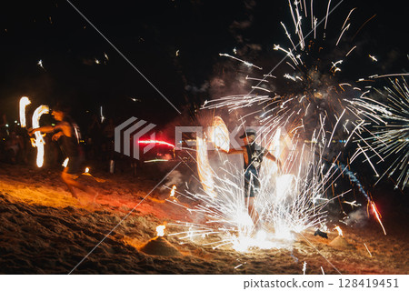 Performers spin fire props, creating light trails, as fireworks burst above a sandy beach. A dark background highlights the fiery spectacle and audience. Performers spin fire props, creating light trails, as fireworks burst above a sandy beach. A dark background highlights the fiery spectacle and audience. 128419451