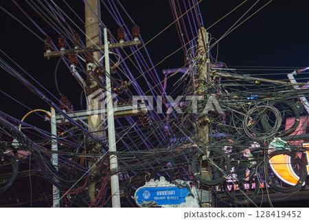 Dense network of electrical wires on utility poles with a blue Bangla Road sign in Thai and English, set against neon lights in Phuket's nightlife area. Dense network of electrical wires on utility poles with a blue Bangla Road sign in Thai and English, set against neon lights in Phuket's nightlife area. 128419452