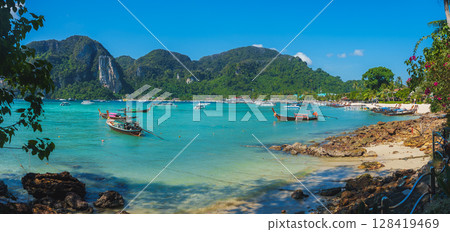 Turquoise waters, lush green cliffs, and anchored boats define this serene Koh Phi Phi beach scene, framed by vibrant foliage and a clear blue sky. 128419469