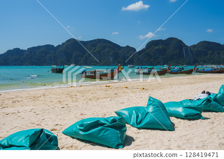 A sandy beach on Koh Phi Phi, Thailand, featuring turquoise waters, limestone cliffs, longtail boats, teal bean bags, and a clear blue sky. 128419471