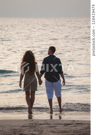 A man and woman walk along a tranquil beach at sunset, with calm ocean waves and soft lighting. Likely set in Phuket or Koh Phi Phi islands, Thailand. 128419474