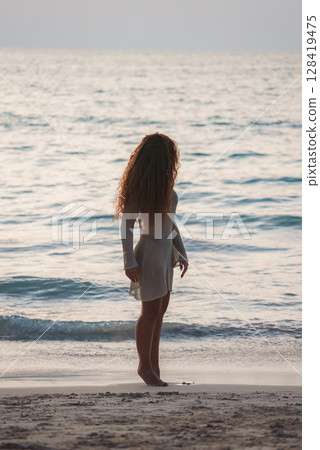 A woman with long curly hair stands barefoot on a sandy beach, wearing a light dress. She faces the calm ocean under soft, warm lighting in Phuket or Koh Phi Phi. A woman with long curly hair stands barefoot on a sandy beach, wearing a light dress. She faces the calm ocean under soft, warm lighting in Phuket or Koh Phi Phi. 128419475