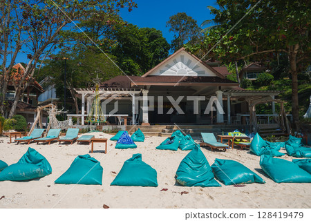 A sandy beach with turquoise bean bags, lounge chairs, and a festive Christmas tree shaped decoration near a small open air building surrounded by greenery. A sandy beach with turquoise bean bags, lounge chairs, and a festive Christmas tree shaped decoration near a small open air building surrounded by greenery. 128419479