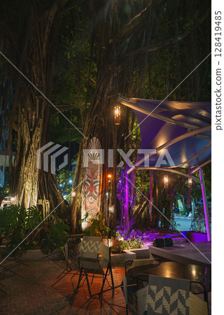 Illuminated banyan trees with hanging lights and purple accents surround modern furniture and a decorative totem in a tropical outdoor setting. 128419485