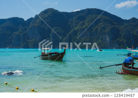 Serene tropical seascape with longtail boats on clear turquoise waters, lush limestone cliffs, and a vibrant blue sky in Koh Phi Phi, Thailand. 128419487