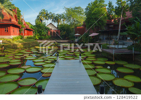 A tranquil pond with green lily pads, a white pier, and a small boat, surrounded by Thai style red wooden buildings and lush tropical greenery. A tranquil pond with green lily pads, a white pier, and a small boat, surrounded by Thai style red wooden buildings and lush tropical greenery. 128419522