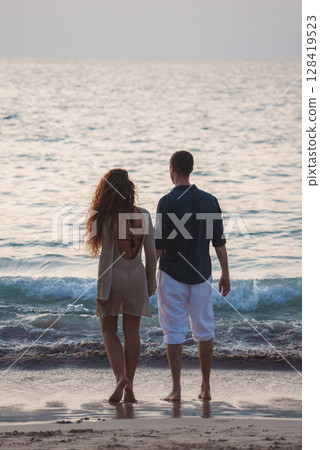 A couple faces the ocean on a sandy beach in Thailand, with soft warm light, gentle waves, and a calm horizon blending with the sea. A couple faces the ocean on a sandy beach in Thailand, with soft warm light, gentle waves, and a calm horizon blending with the sea. 128419523