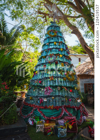 A festive Christmas tree made from green plastic bottles with ribbons and ornaments, set outdoors among palm trees and tropical greenery in Thailand. A festive Christmas tree made from green plastic bottles with ribbons and ornaments, set outdoors among palm trees and tropical greenery in Thailand. 128419537