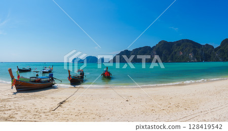 A serene beach in Koh Phi Phi, Thailand, featuring turquoise waters, white sand, longtail boats with colorful ribbons, and dramatic limestone cliffs. A serene beach in Koh Phi Phi, Thailand, featuring turquoise waters, white sand, longtail boats with colorful ribbons, and dramatic limestone cliffs. 128419542