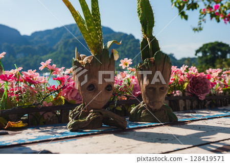Two Groot shaped plant pots with small plants on a wooden surface, surrounded by pink and red flowers, with green hills and blue sky in the background. Two Groot shaped plant pots with small plants on a wooden surface, surrounded by pink and red flowers, with green hills and blue sky in the background. 128419571