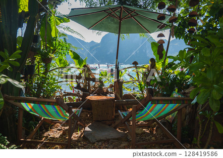 Tropical seating area with striped deck chairs, green umbrella, and wooden table, overlooking boats and limestone cliffs in Koh Phi Phi, Thailand. Tropical seating area with striped deck chairs, green umbrella, and wooden table, overlooking boats and limestone cliffs in Koh Phi Phi, Thailand. 128419586