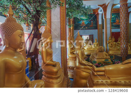 Richly adorned temple interior in Thailand featuring golden Buddha statues, intricate gold and red columns, vibrant murals, and a decorative tree. 128419608