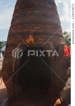 A large, conical brick kiln with a dark cavity at its base, adorned with a marigold garlanded statue, surrounded by trees and structures in Thailand. 128419611