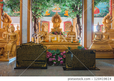 A golden reclining Buddha statue surrounded by seated Buddhas, vibrant murals, decorative trees, and black plaques with gold inscriptions in a Thai temple. 128419614