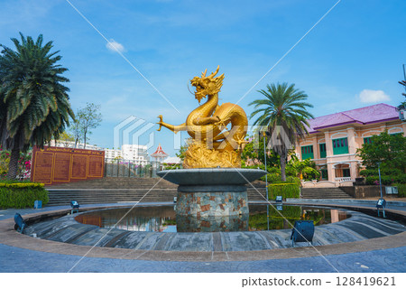 Golden dragon statue atop a circular fountain surrounded by gardens, palm trees, and a pink roofed building under a clear blue sky in Thailand. Golden dragon statue atop a circular fountain surrounded by gardens, palm trees, and a pink roofed building under a clear blue sky in Thailand. 128419621
