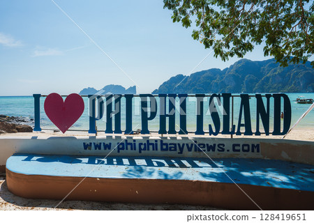 A sign reading 'I PHI PHI ISLAND' stands on a sandy beach with turquoise waters, limestone cliffs, and tree branches in Koh Phi Phi, Thailand. 128419651