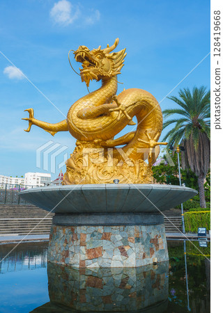 Golden dragon statue with coiled body and open mouth on a circular stone pedestal, surrounded by water, greenery, and a clear blue sky in Phuket. Golden dragon statue with coiled body and open mouth on a circular stone pedestal, surrounded by water, greenery, and a clear blue sky in Phuket. 128419668