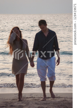 A man and woman walk hand in hand along a sandy beach at sunset, with the ocean shimmering in the background. Likely in Phuket or Koh Phi Phi islands. 128419671