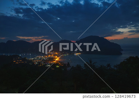 Koh Phi Phi at night with town lights reflecting on calm bay waters, lush vegetation in the foreground, and limestone cliffs in the background. 128419673