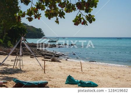 A tranquil beach in Phuket, Thailand, with golden sand, turquoise waters, long tail boats, a tripod structure, and shaded teal bean bags under trees. A tranquil beach in Phuket, Thailand, with golden sand, turquoise waters, long tail boats, a tripod structure, and shaded teal bean bags under trees. 128419682