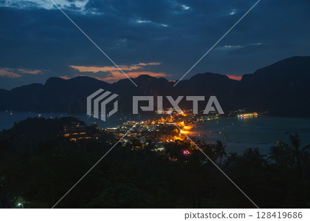 Koh Phi Phi's vibrant lights reflect on the water, surrounded by dark limestone cliffs and lush vegetation under a deep blue and orange sky. 128419686