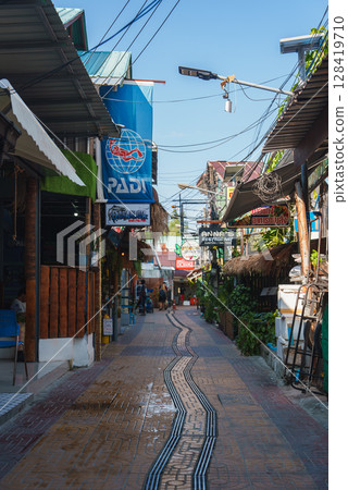 A colorful street in Thailand featuring shops, a PADI diving center, Anna's Restaurant, tiled pathways, greenery, and overhead electrical wires. 128419710
