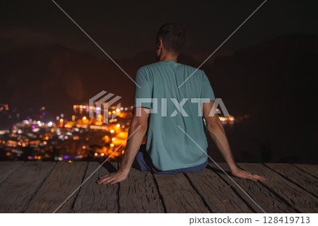 A man in a light blue shirt sits on a wooden platform at night, gazing at a brightly lit cityscape below, surrounded by dark hills in Thailand. 128419713