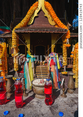A small Thai spirit house adorned with marigold garlands, colorful fabric, and offerings like red liquid bottles, incense, and figurines under bright sunlight. 128419743