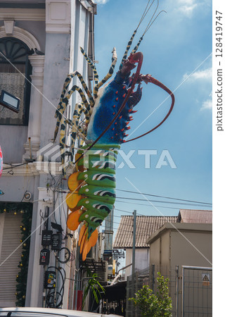 Large lobster sculpture in vibrant colors decorates a white building with arched windows, set in an urban area with tropical plants and a blue sky. Large lobster sculpture in vibrant colors decorates a white building with arched windows, set in an urban area with tropical plants and a blue sky. 128419747