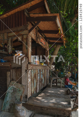 A weathered wooden shack with a corrugated metal roof surrounded by lush greenery, featuring a small counter with items and a seated person nearby. 128419749
