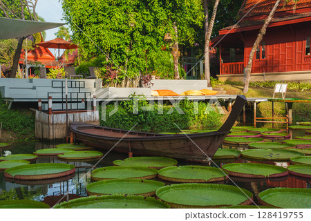 A wooden Thai boat rests on a pond with green lily pads, surrounded by red roofed Thai style buildings, trees, and a modern seating area in Thailand. A wooden Thai boat rests on a pond with green lily pads, surrounded by red roofed Thai style buildings, trees, and a modern seating area in Thailand. 128419755