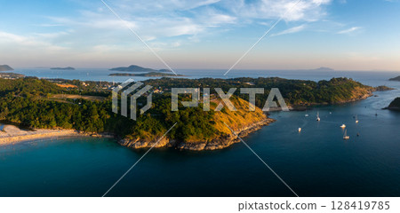 Aerial view of Naiharn Beach in Phuket, Thailand, featuring golden sands, turquoise waters, anchored sailboats, lush hills, and a distant lighthouse. 128419785