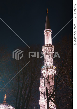 A brightly lit minaret with intricate details and a golden finial stands against a dark night sky, partially framed by bare tree branches in Istanbul, Turkey. 128419797