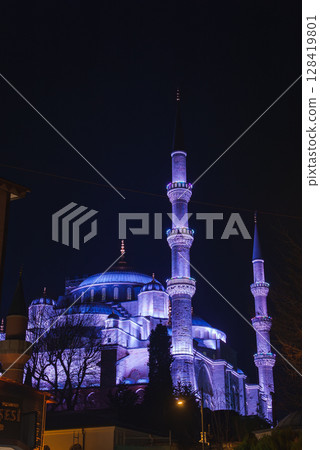 The Sultan Ahmed Mosque, with its domes and six minarets lit in purple and blue, stands against a dark sky, framed by trees and nearby structures. 128419801