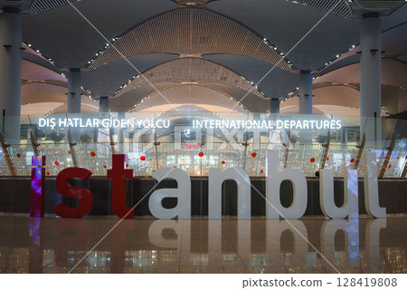 The interior of Istanbul Airport features a bold illuminated Istanbul sign, modern arched ceilings, sleek columns, and reflective flooring near departures. 128419808