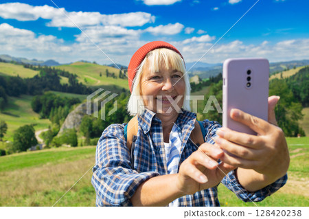 Smiling senior woman taking a selfie during a hiking adventure in the mountains 128420238
