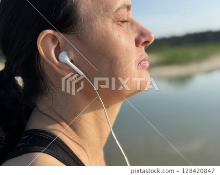 Close up adult sporty woman relaxing, listening to music in headphones after workout by lake in forest. Outdoor fitness, recovery, peaceful summer nature scene Close up adult sporty woman relaxing, listening to music in headphones after workout by lake in forest. Outdoor fitness, recovery, peaceful summer nature scene 128420847