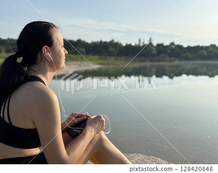 Close up adult sporty woman relaxing, listening to music in headphones after workout by lake in forest. Outdoor fitness, recovery, peaceful summer nature scene 128420848