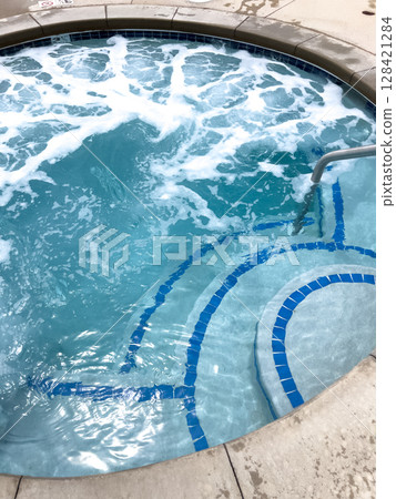 A round hot tub filled with bubbling water shows blue tile patterns and curved steps. A handrail extends into the water under bright lighting. A round hot tub filled with bubbling water shows blue tile patterns and curved steps. A handrail extends into the water under bright lighting. 128421284